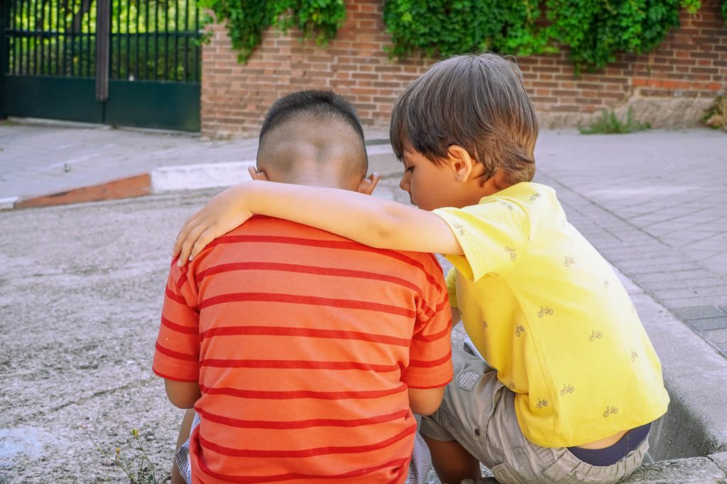Dirljiv trenutak dečije empatije - dečak u žutoj majici teši svog druga, pokazujući iskreno prijateljstvo i emocionalnu podršku kroz spontani zagrljaj.